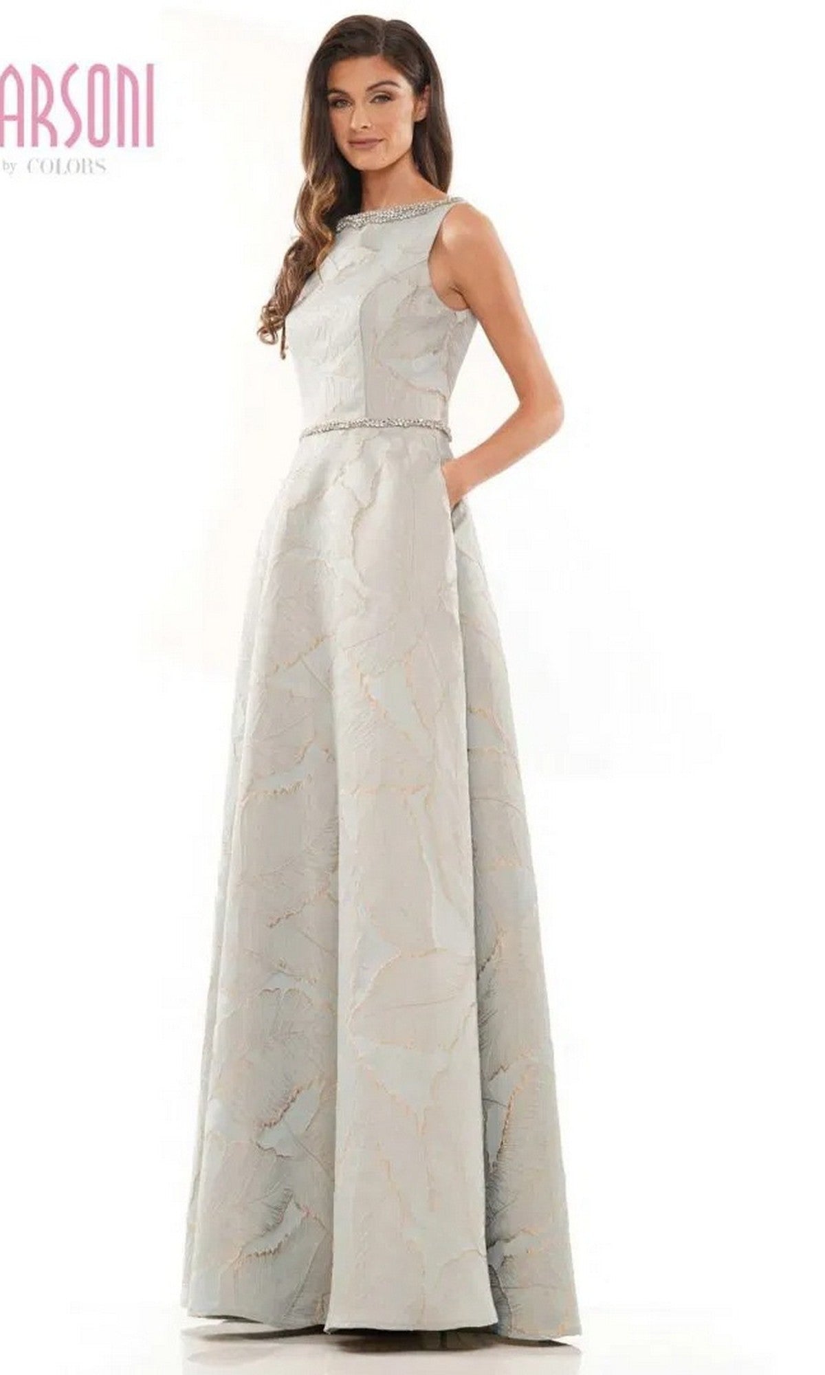 A woman wears the Marsoni MV1224 long formal dress in seaglass, featuring a floor-length, sleeveless design with a subtle jacquard print and jeweled waistband, standing with one hand in her pocket.